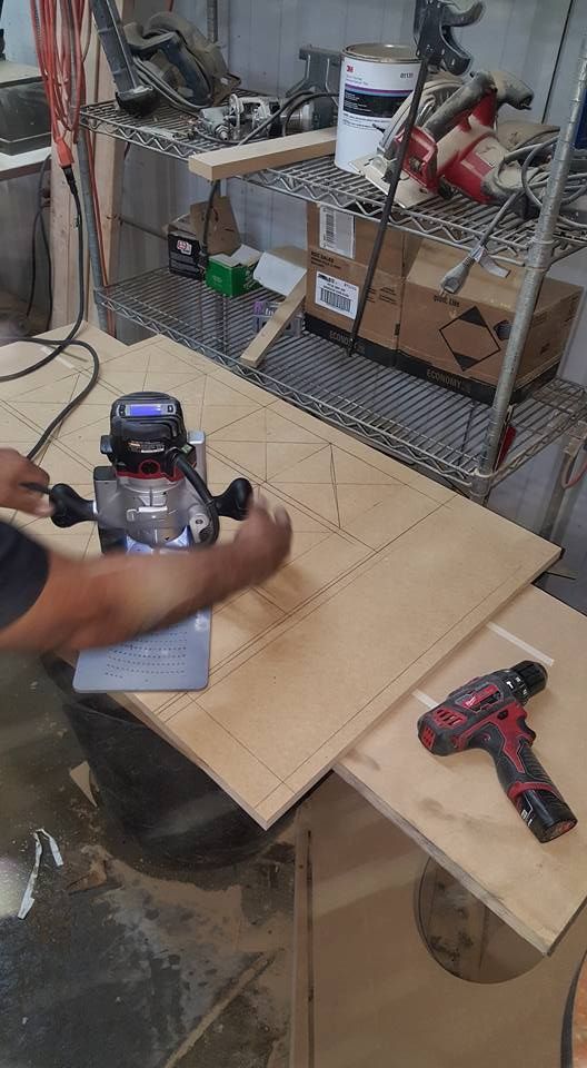 A person using a router to cut a design in a wood panel. The workshop setting includes tools and supplies.