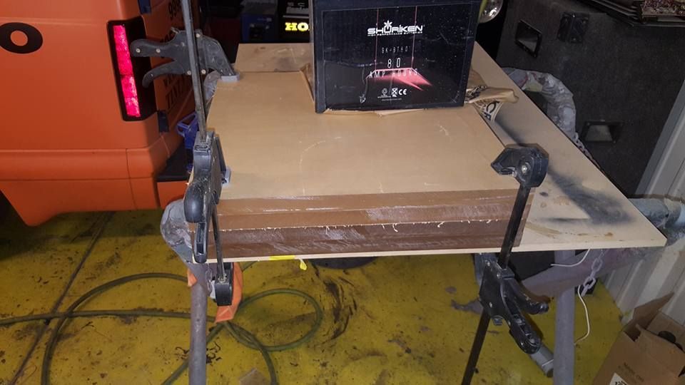 A wooden surface clamped together, with a black device on top, possibly a speaker. Orange vehicle visible in the background.