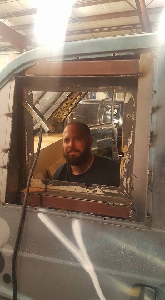 Man with a beard looking through a rectangular opening in a vehicle's side; he's in a shop.