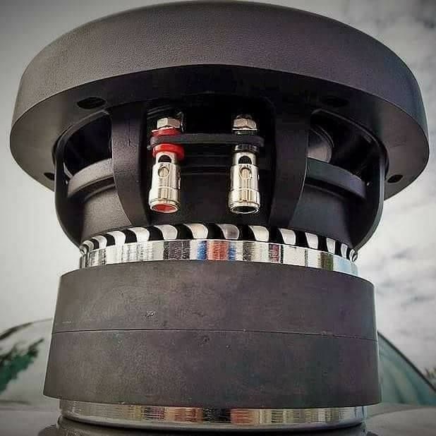 Close-up of a black subwoofer with red and silver speaker terminals. It's mounted, with a car visible in the background.