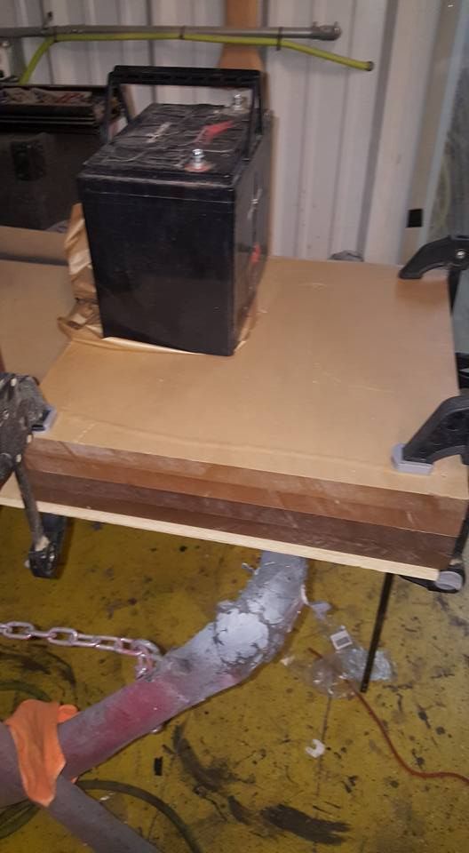 A car battery sits on a stack of cardboard sheets clamped to a surface in a workshop.