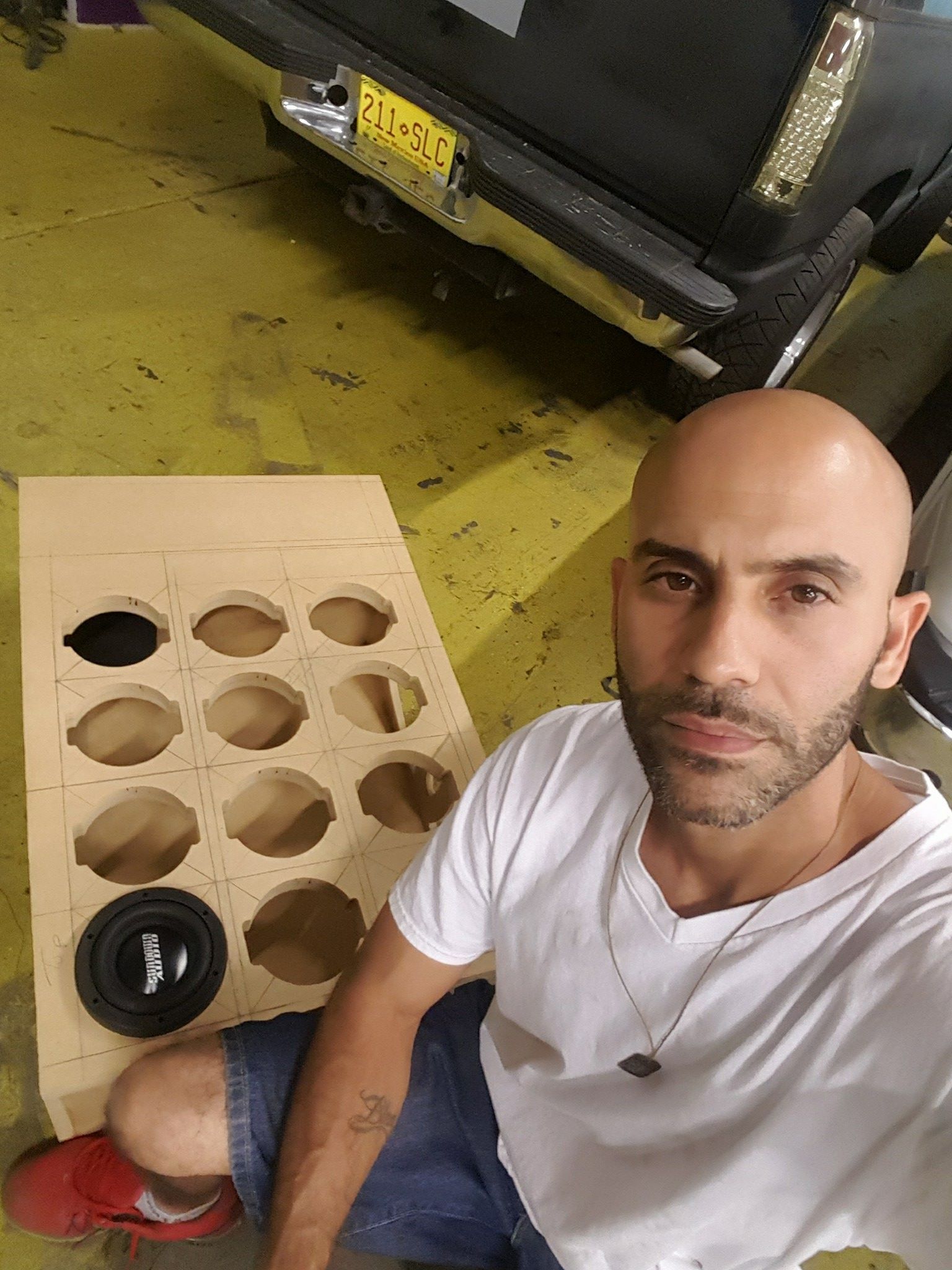 Bald man with a light complexion sits beside a wooden board with circular holes, preparing to install a car speaker.