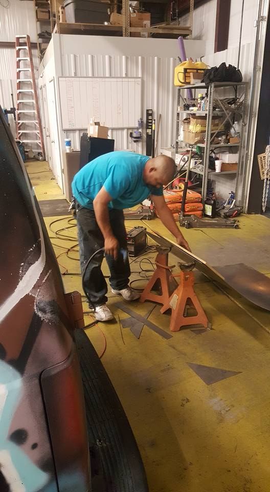 Man in blue shirt works with metal sheet supported by jack stands in a workshop with tools and a ladder.