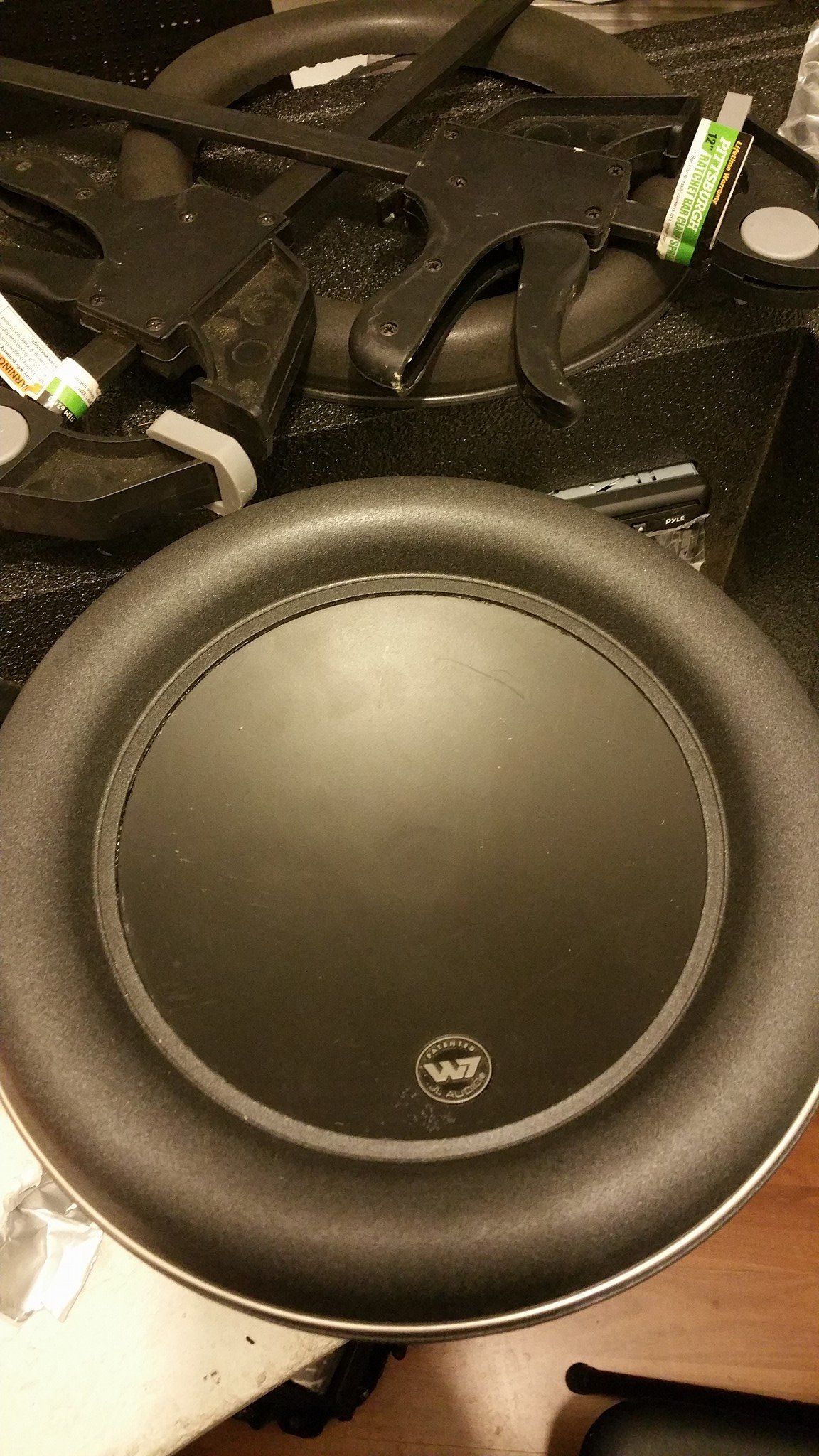 Black subwoofer speaker with a thick rubber surround on a cluttered surface.