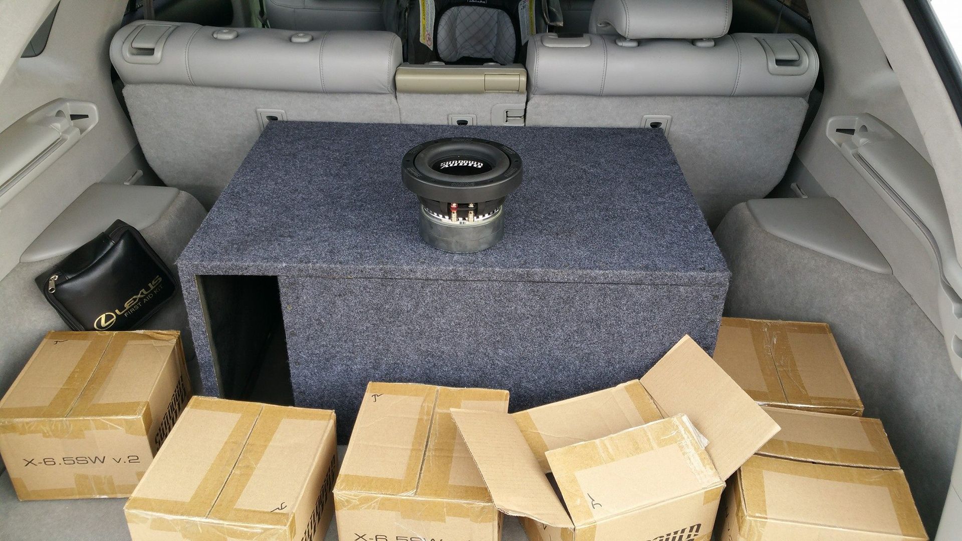Gray car trunk filled with boxes and a subwoofer box with a subwoofer on top.