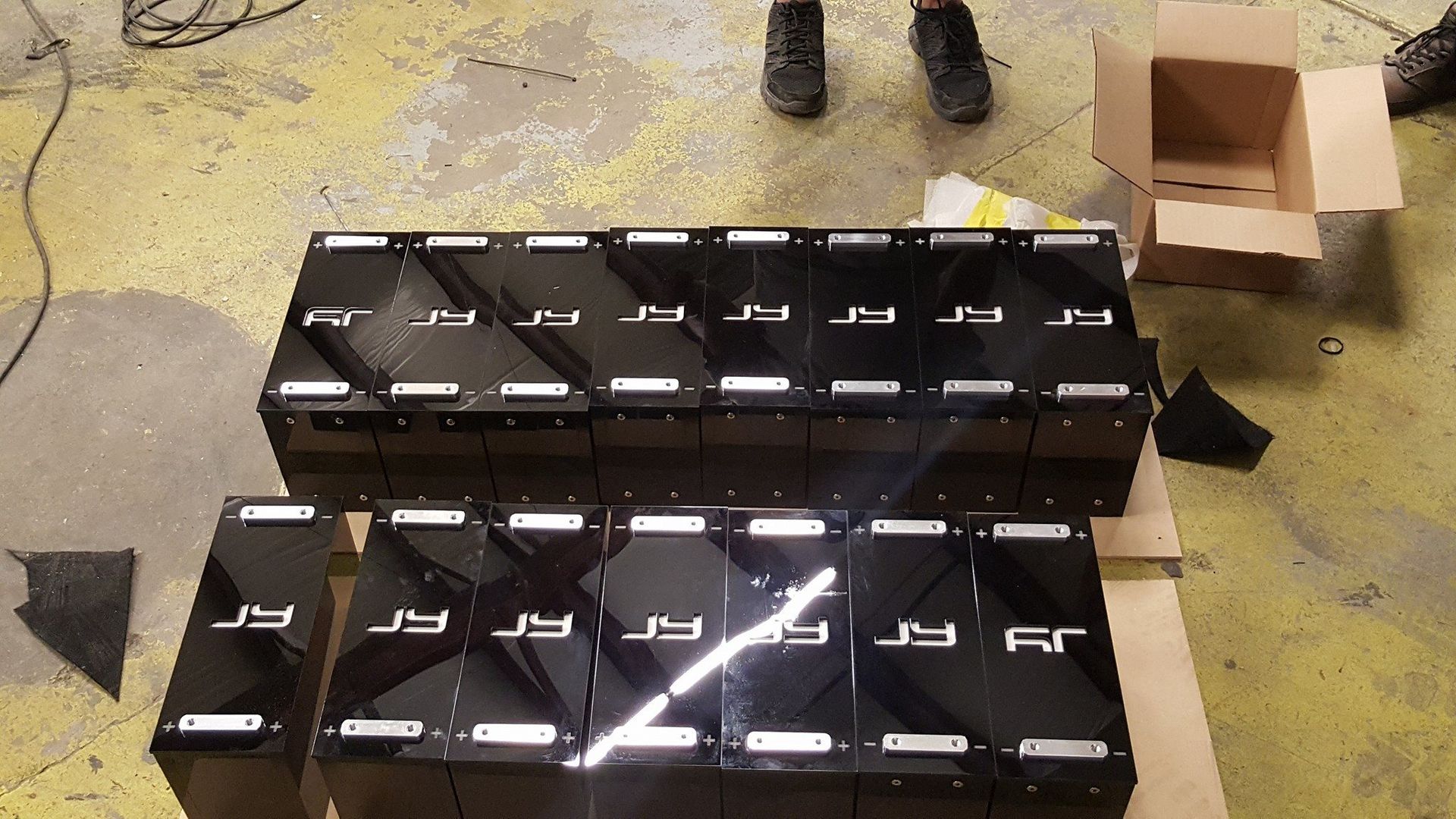 Black battery packs stacked on a wooden pallet, with a person's legs and cardboard box in the background.