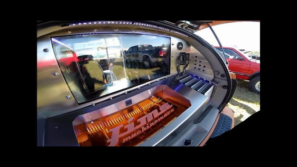 Interior of a custom vehicle, featuring a large display screen and intricate lighting; visible in the reflection is a truck.