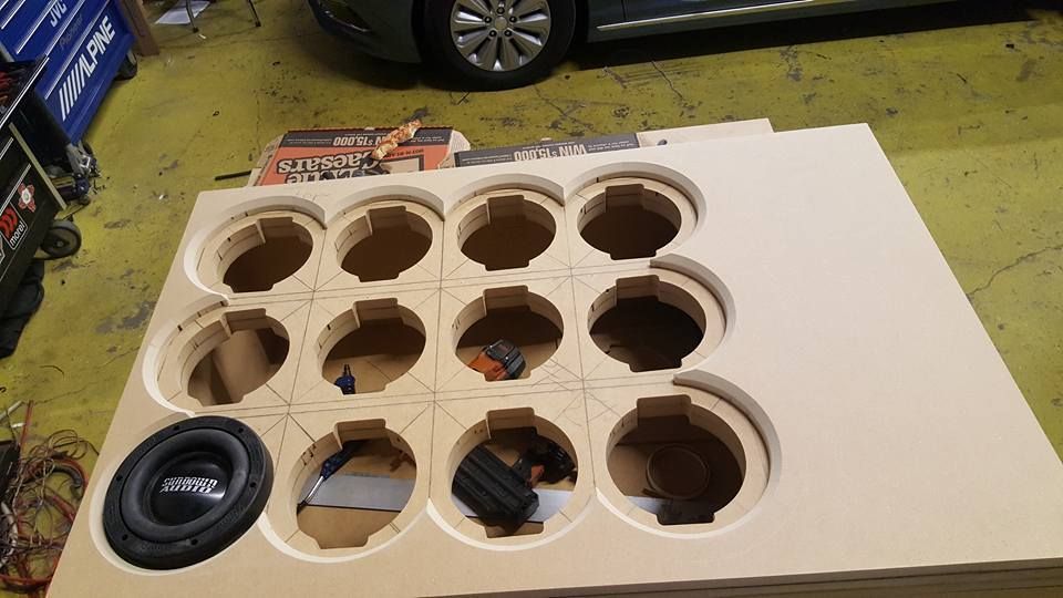A large unfinished speaker box with many round cutouts. A subwoofer is installed in one of the holes.