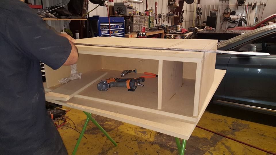 A person builds a wooden structure with compartments inside, possibly for a car's audio system, in a workshop.