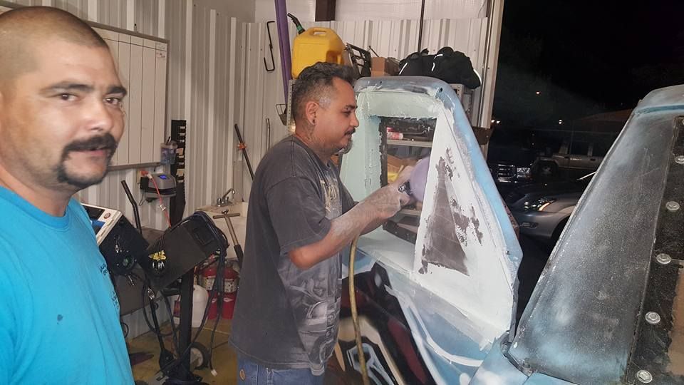 Two men working on a car body in a garage. One grinds, the other looks at the camera.