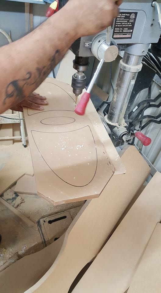 Person using a drill press to bore holes in a piece of wood with a traced design in a workshop.