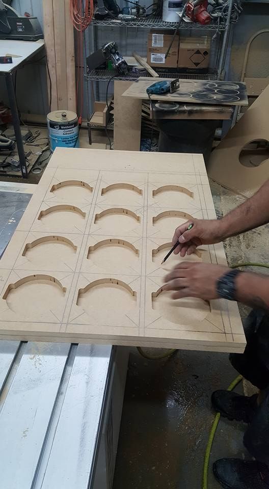Person tracing a template on a large wood panel with pre-cut circular and rectangular outlines in a workshop setting.
