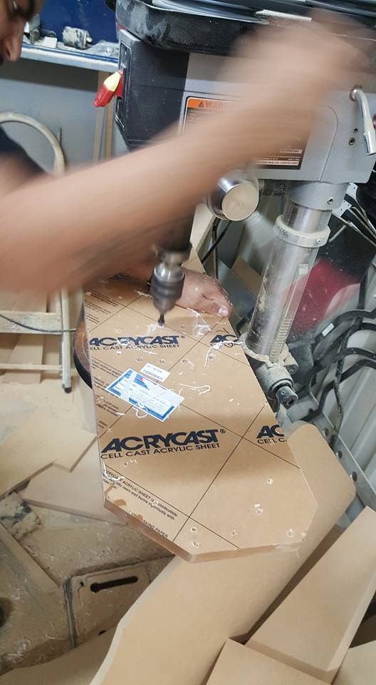 A person drills into a piece of clear acrylic with a drill press in a workshop. Sawdust is visible.