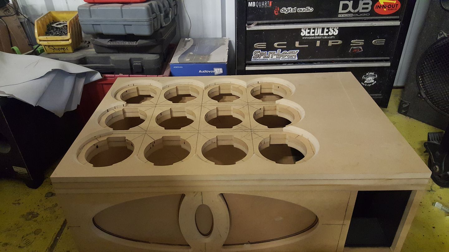 A wooden speaker enclosure with twelve circular cutouts for speakers and a decorative oval design.
