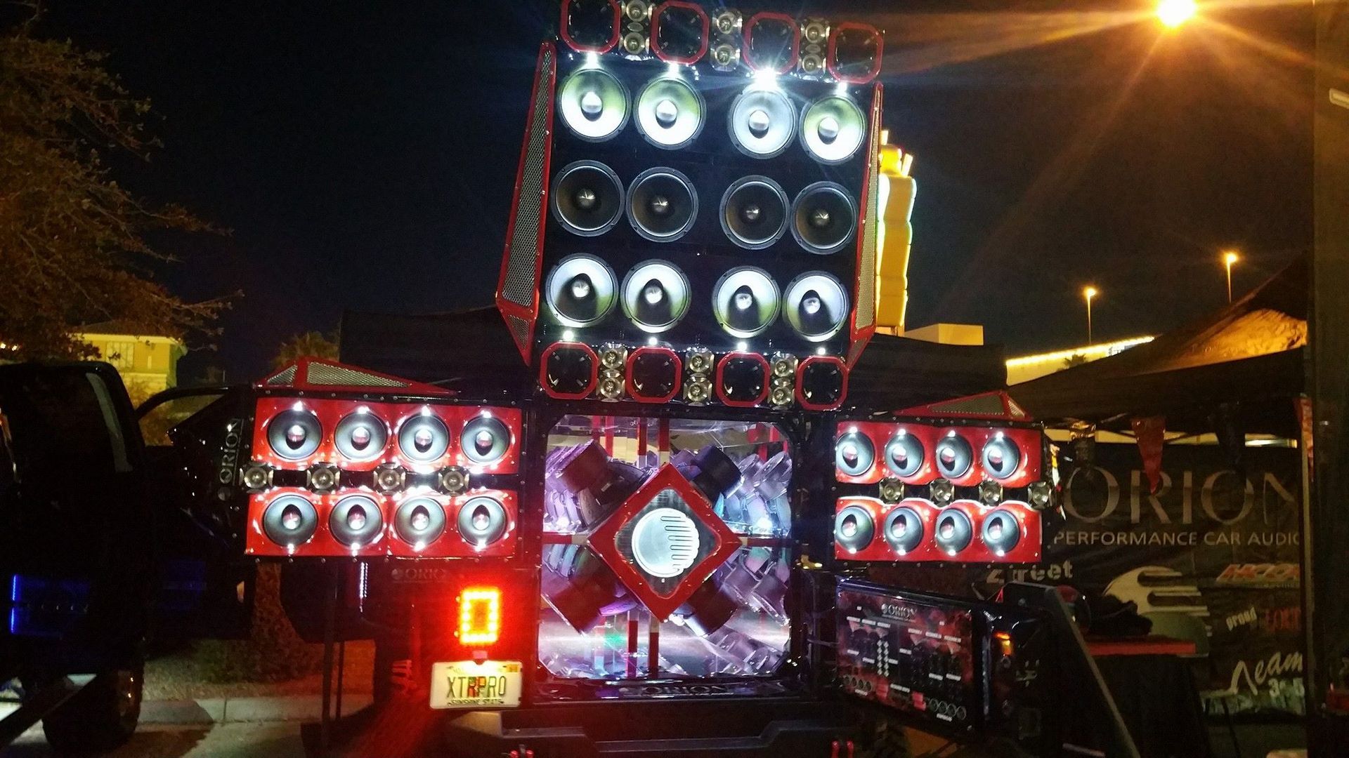 A truck bed overflowing with speakers, lit up at night with red and white lights; the Orion logo is partially visible.