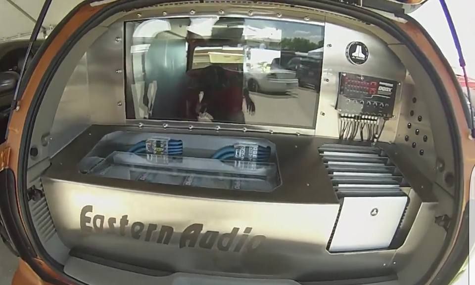 Custom car audio installation in a vehicle's trunk; features a large screen, amplifiers, and the words 