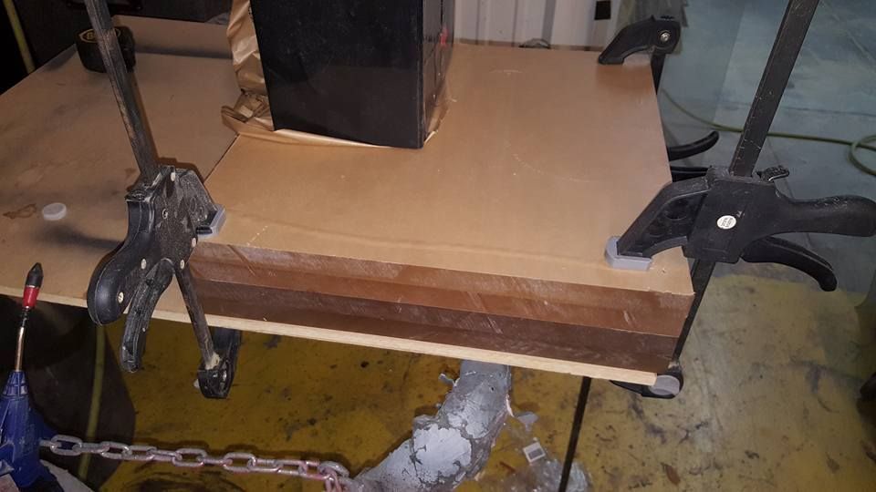 Brown wooden planks held together by clamps, with a dark box on top, in a workshop setting.