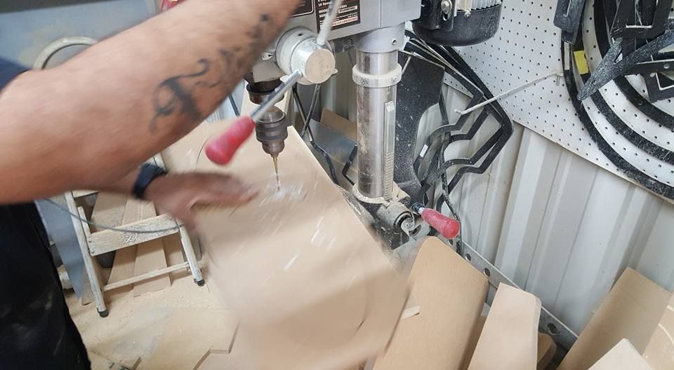 Person using a drill press on a piece of wood in a workshop. Sawdust is visible.