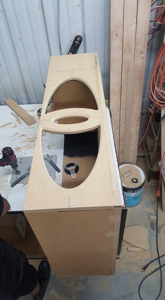Wooden cabinet under construction with oval cutouts for a sink and faucet, on a worktable in a workshop.