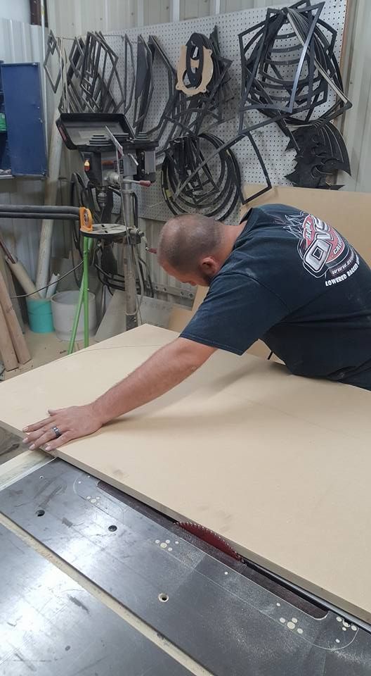 Man cutting a large sheet of wood with a table saw in a workshop. He is wearing a dark shirt and focused on his work.