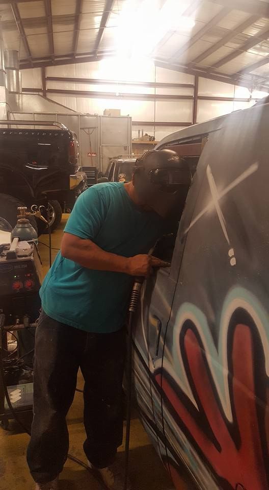 A person wearing a welding mask and blue shirt works on a vehicle with graffiti in a garage.