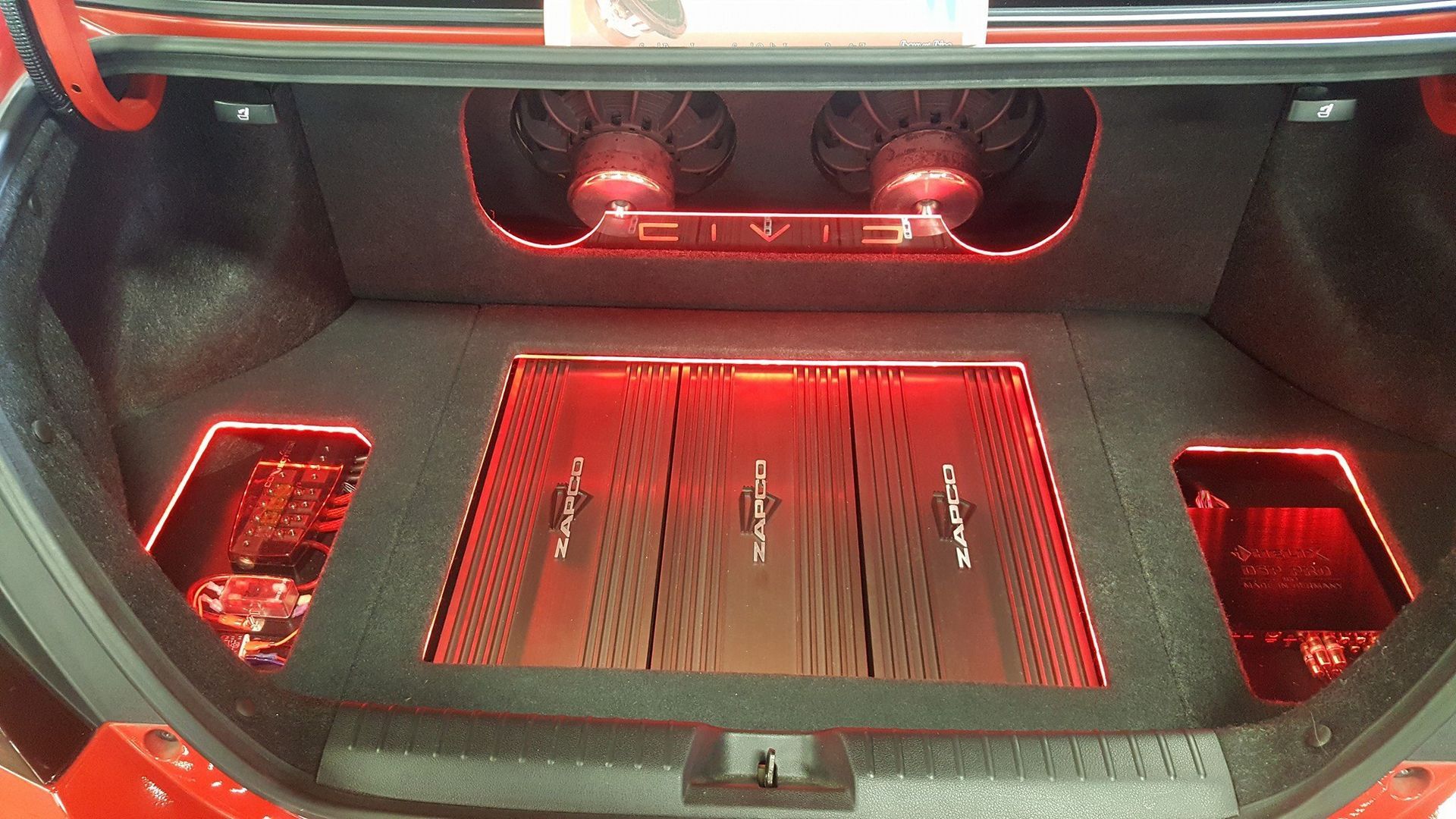 Red-lit car trunk showcasing a custom audio system with speakers and amplifiers against a black interior.