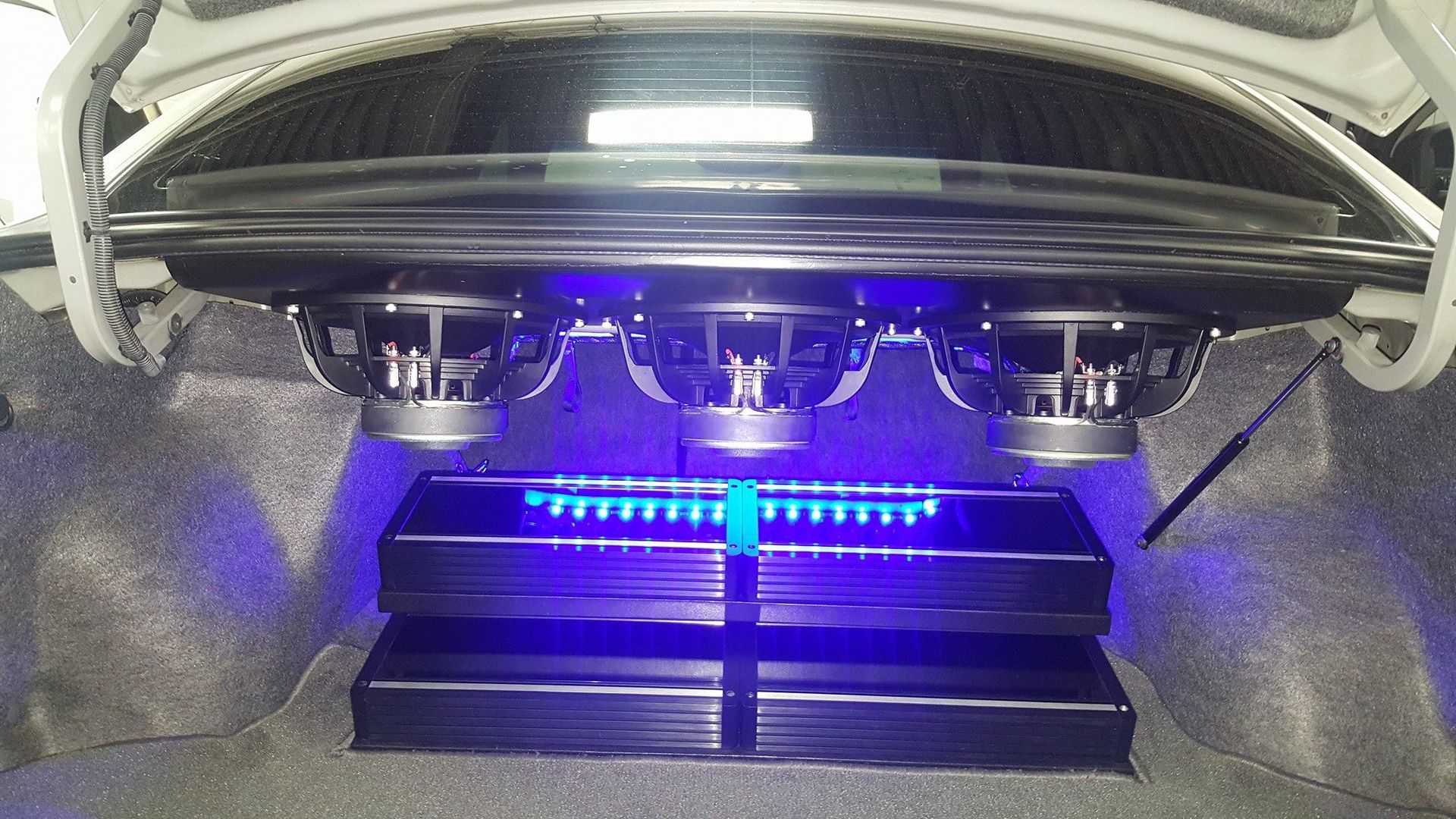 Inside of a car trunk: three large subwoofers, two amplifiers, and blue LED lighting.