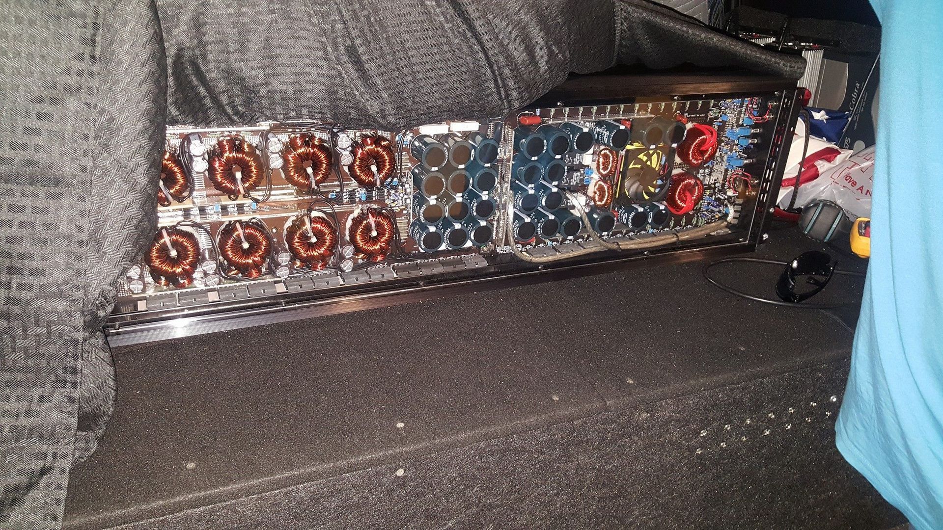 Two car audio amplifiers with exposed circuitry sit in a vehicle's trunk. 