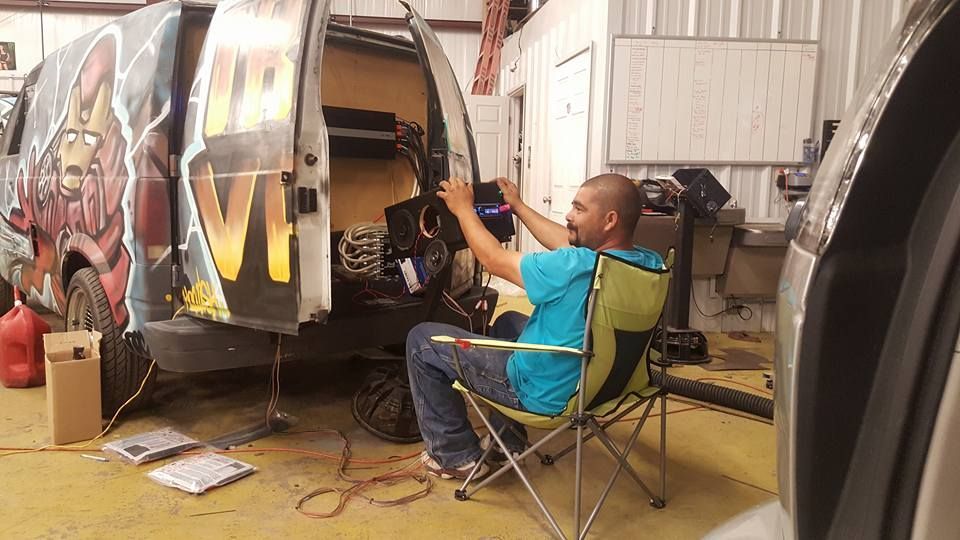 A man in a blue shirt sits in a folding chair, working on the wiring of a van in a garage. The van has graffiti art.
