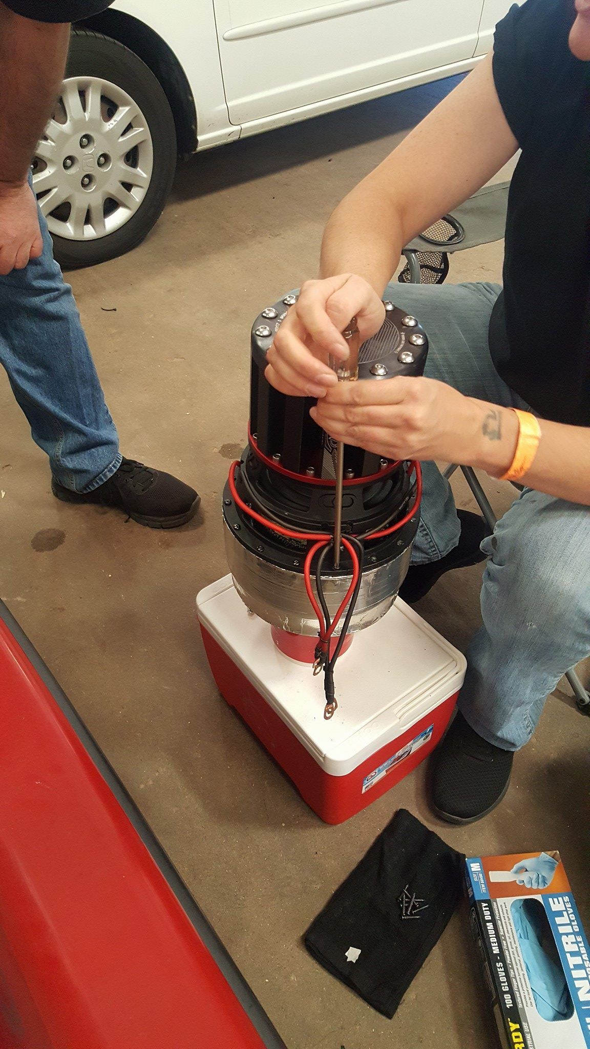 Person working on a large, black cylindrical device, likely mechanical, resting on a cooler, with parts and tools visible.