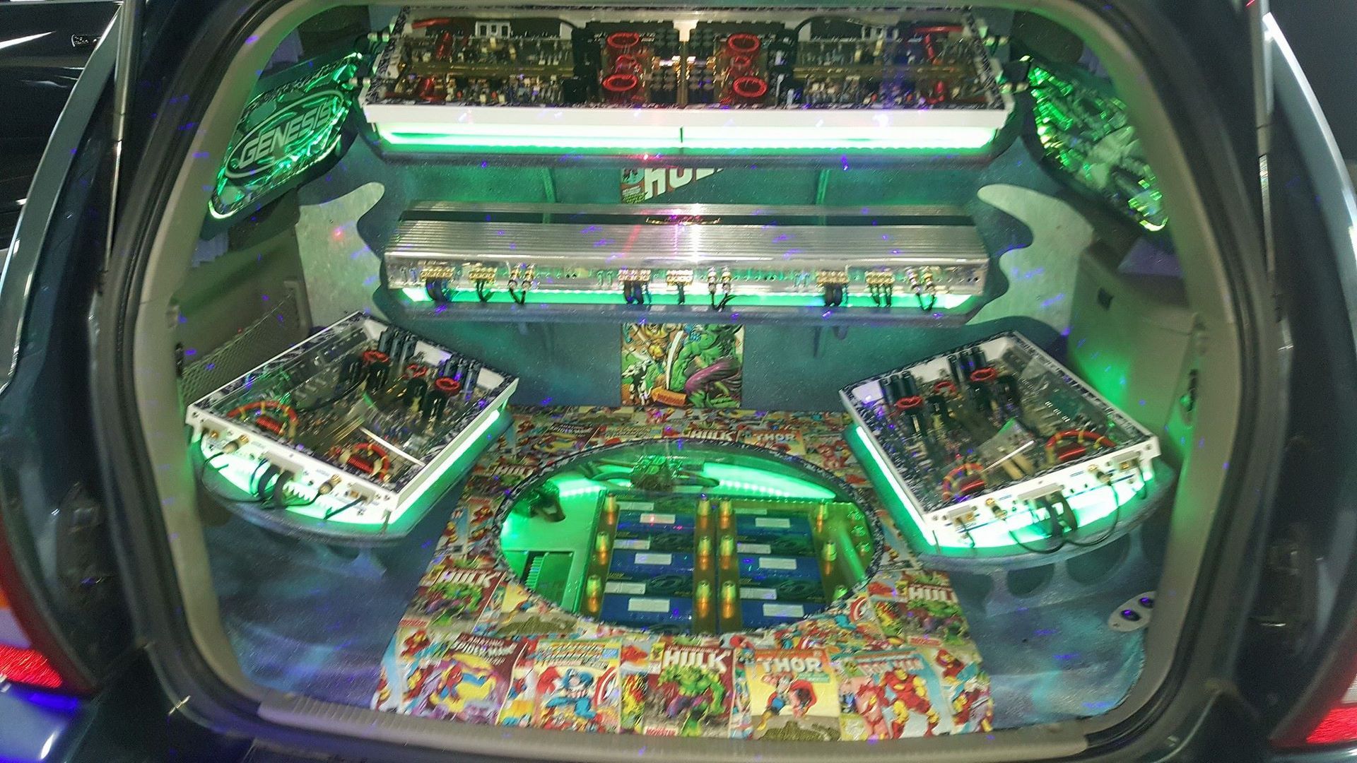 A car trunk customized with multiple amplifiers and lighting, displaying a vibrant green and multi-colored setup.