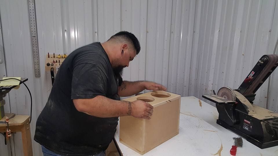 A man assembling a wooden box with cup holders in a workshop. He's wearing a gray shirt and has a beard.
