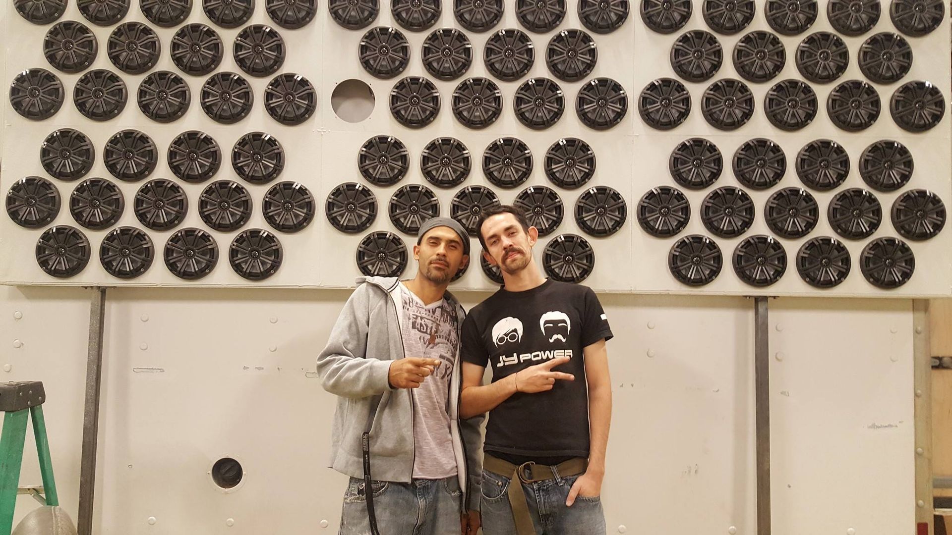Two men pose in front of a wall covered with round, speaker-like objects.