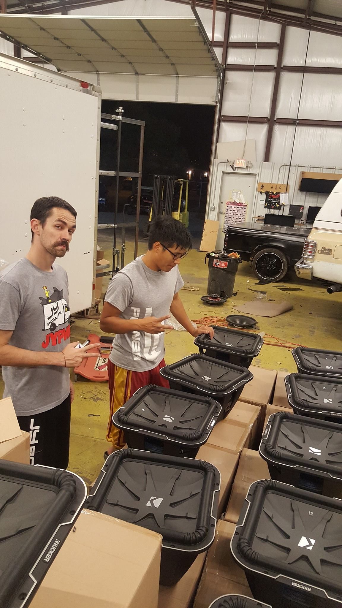 Two men in a warehouse inspecting many black containers stacked on boxes. One man points while the other looks on.