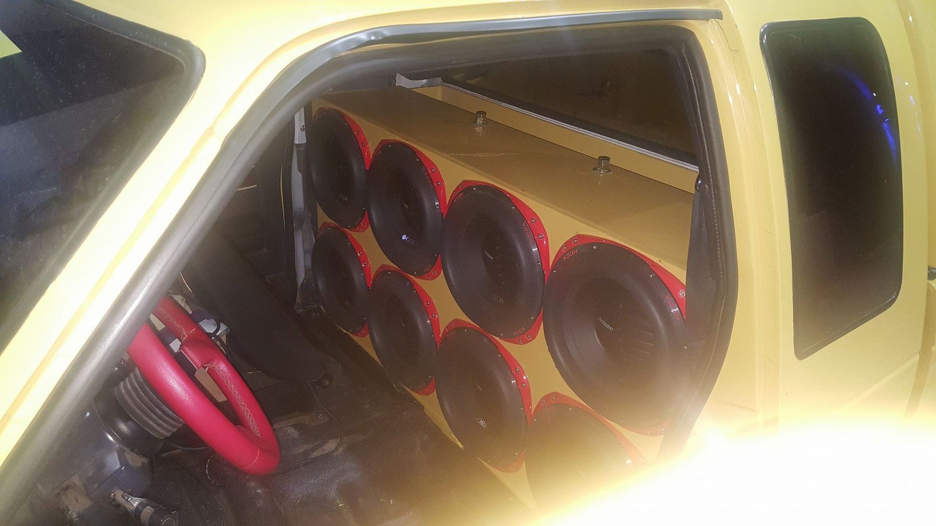 Inside a yellow car: a wall of black subwoofers with red accents fills the back seat area. A red steering wheel is visible.