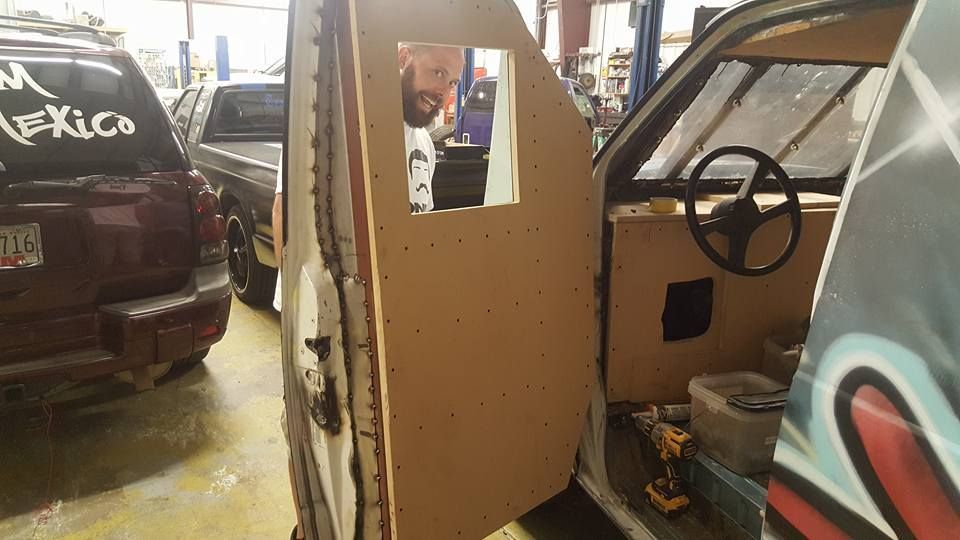 A person looks through a square window in a wooden door, part of a vehicle undergoing modifications in a garage.