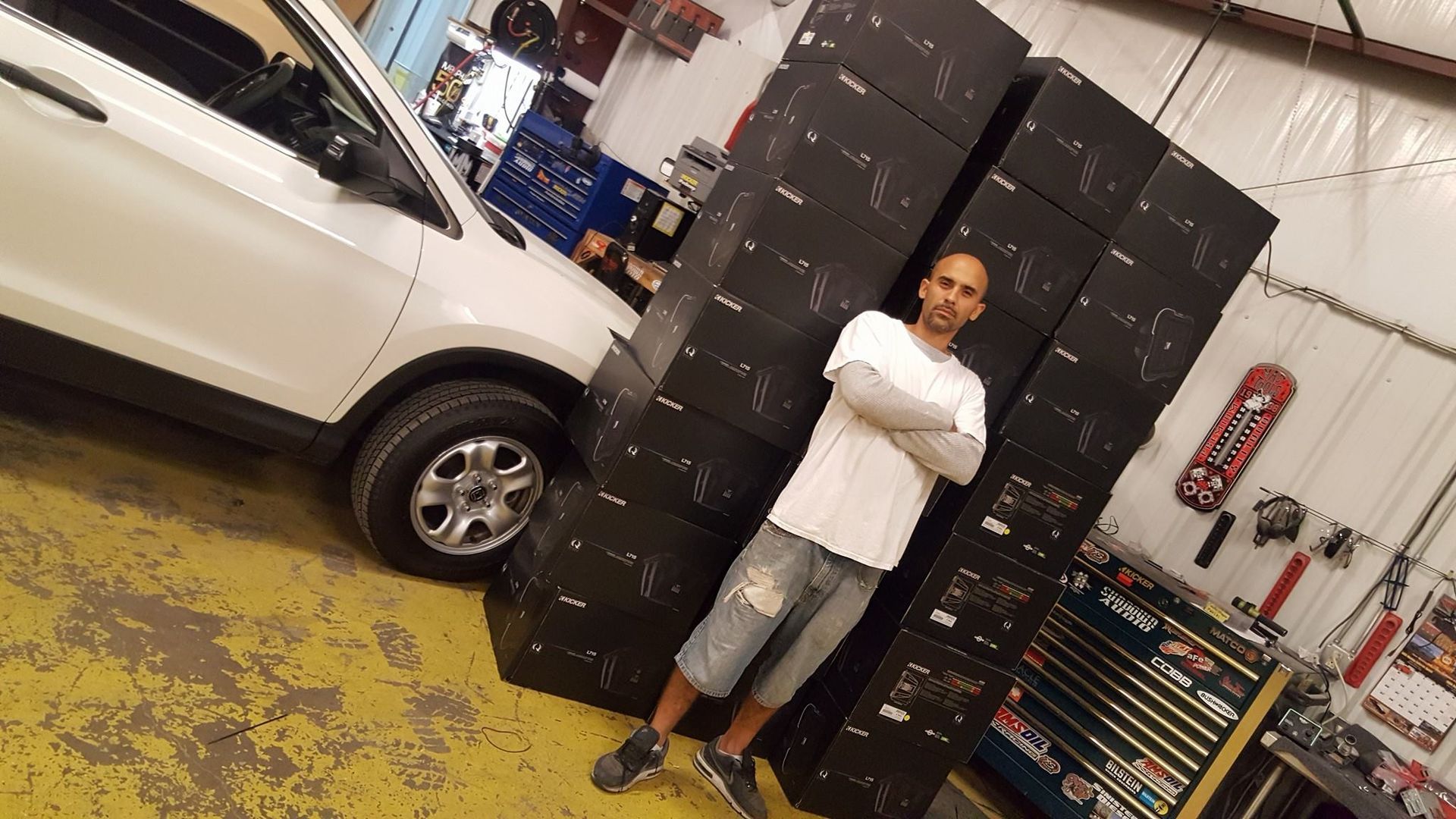 Man standing with arms crossed next to a large stack of black boxes and a white car in a garage.