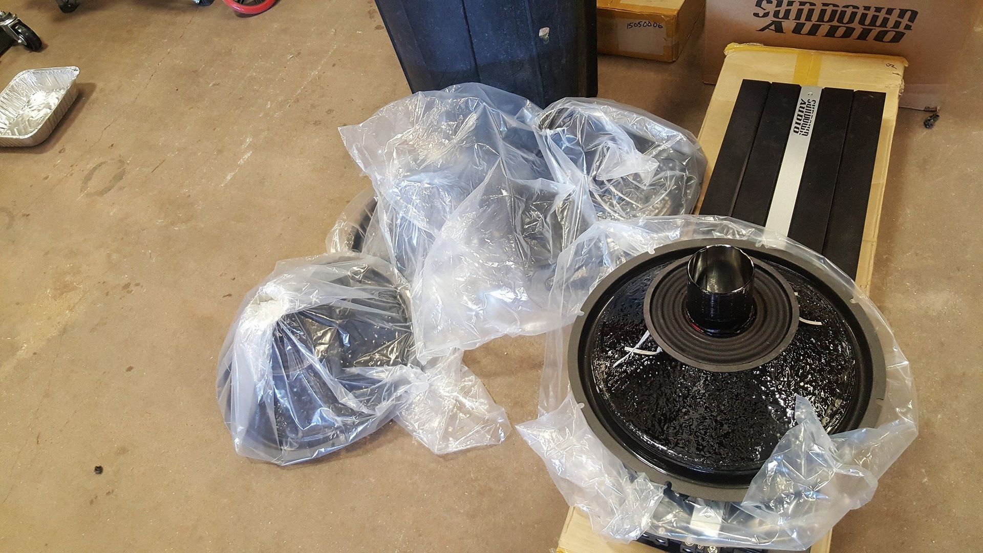 Several black speakers and related components, wrapped in plastic, are lying on a wooden floor.