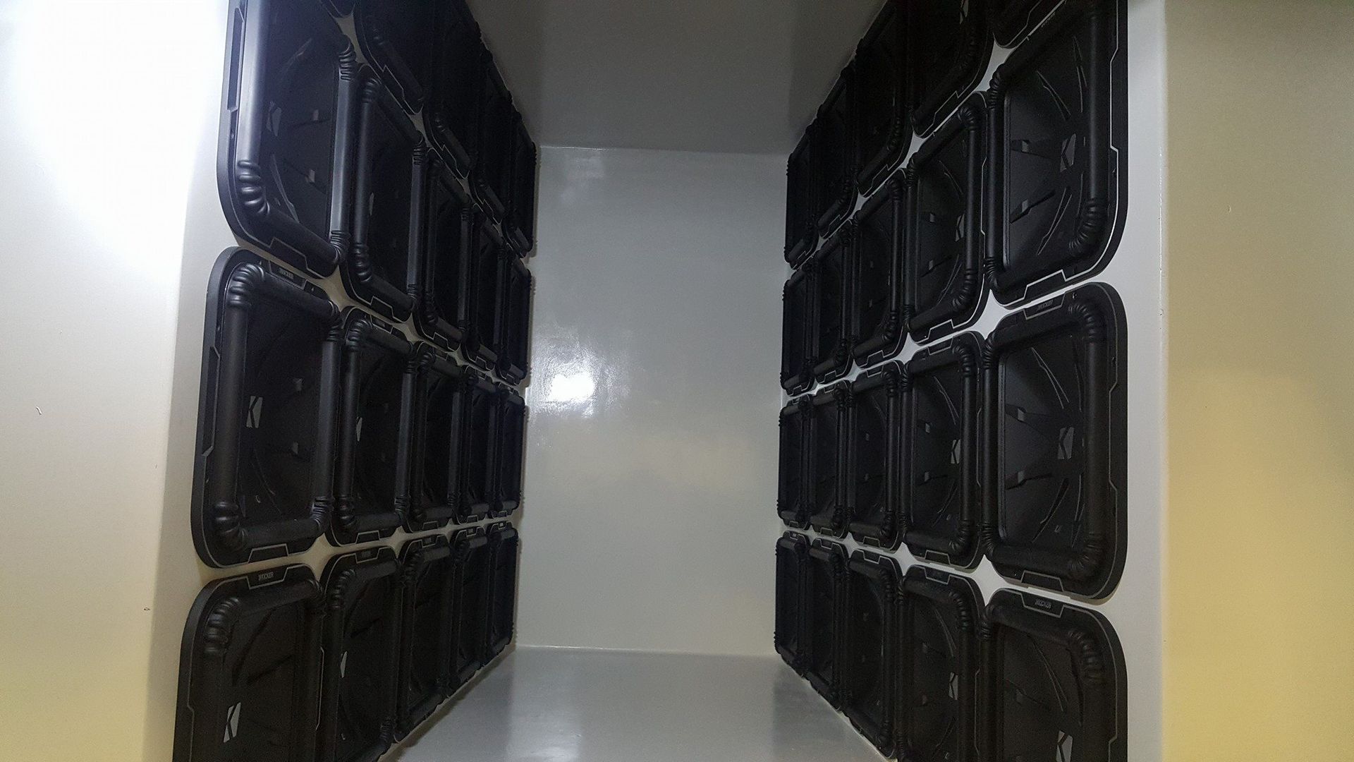 A narrow, white-walled corridor lined with rows of black, rectangular containers. The space suggests storage or containment.