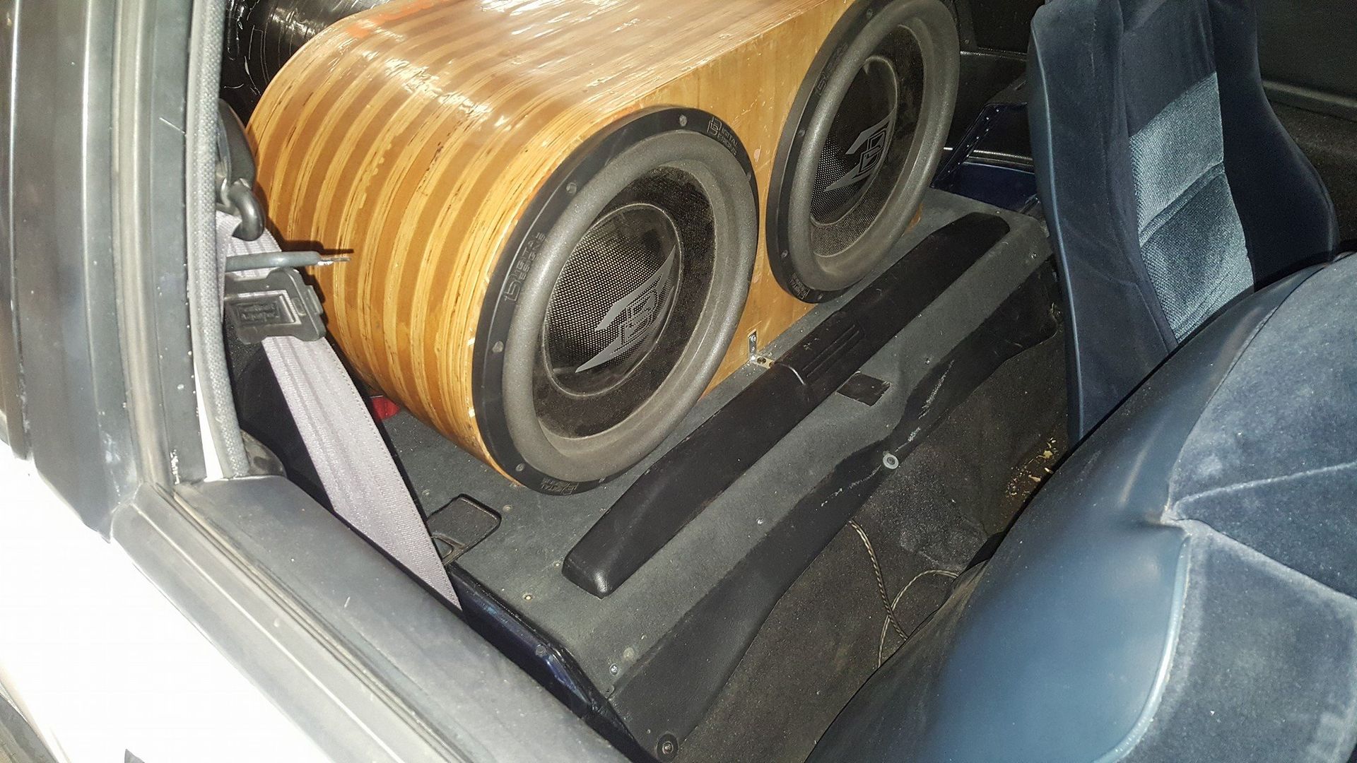 Two subwoofers are mounted in a wooden enclosure in the backseat of a car. The seat is blue and the car interior is dark.