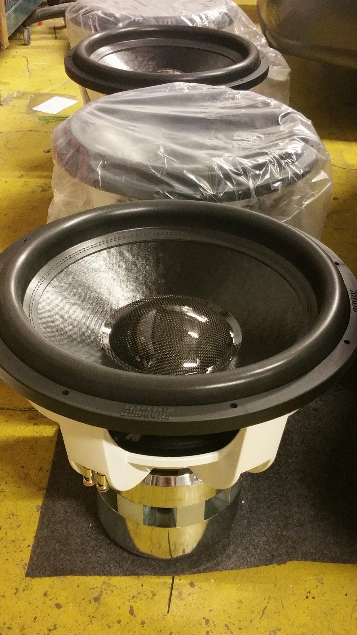 Three large car audio subwoofers, one uncovered, are in a factory setting. 