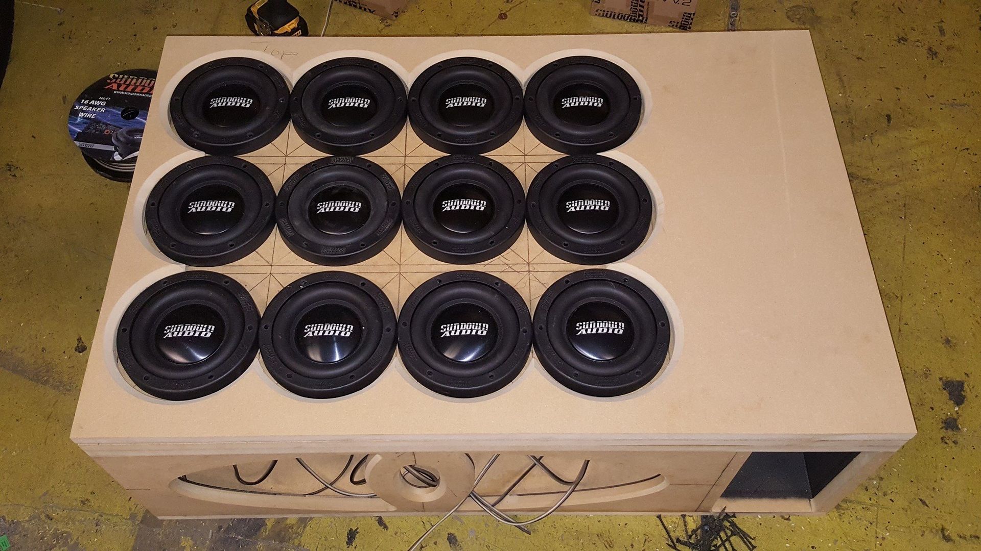 A large wooden subwoofer box with twelve installed speakers. The box is beige and has a decorative design on the front.