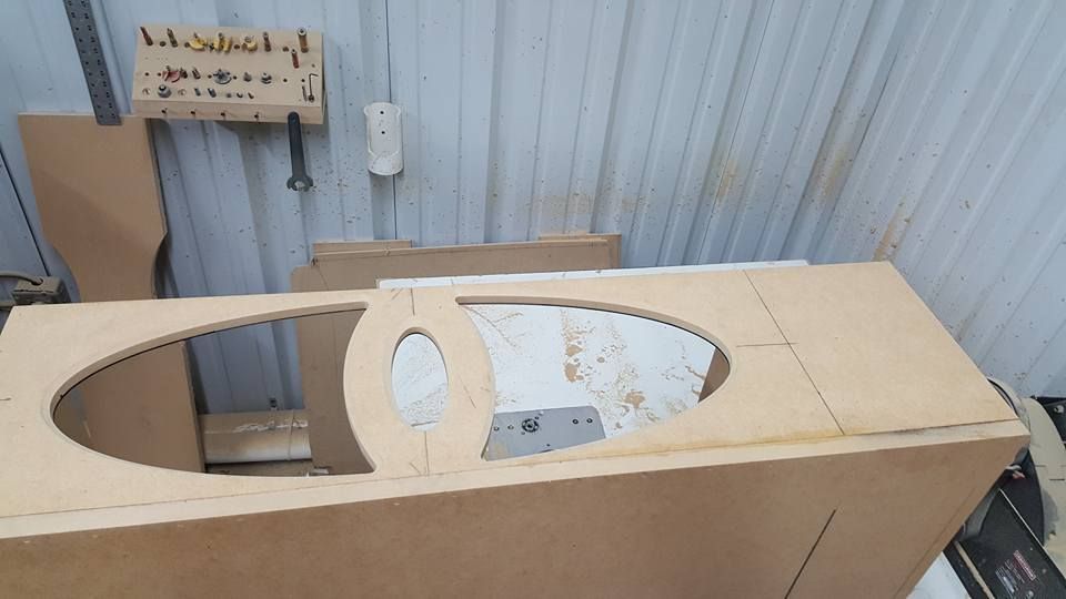 A partially constructed wooden cabinet with oval and circular cutouts sits in a workshop. Beige and brown wood.