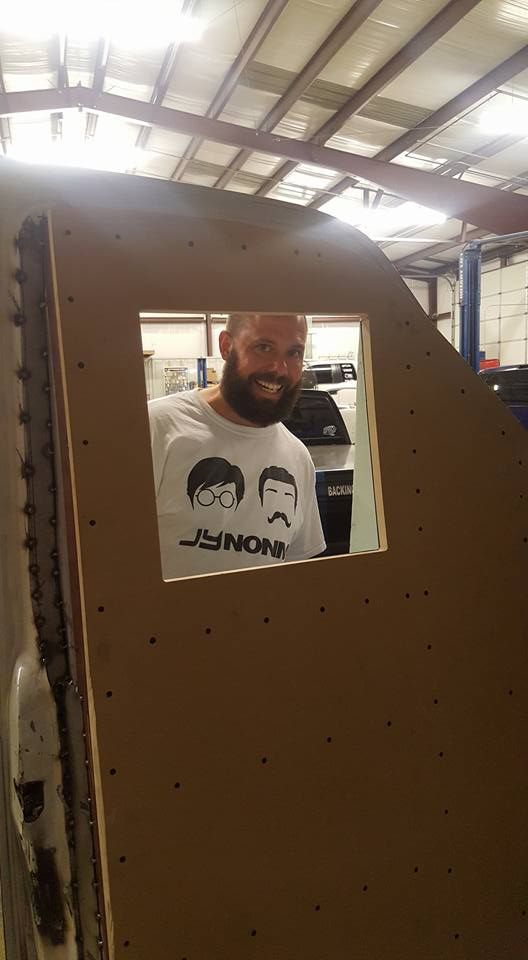 Man with a beard smiles through a square opening in a brown structure, wearing a Beatles t-shirt in a workshop.