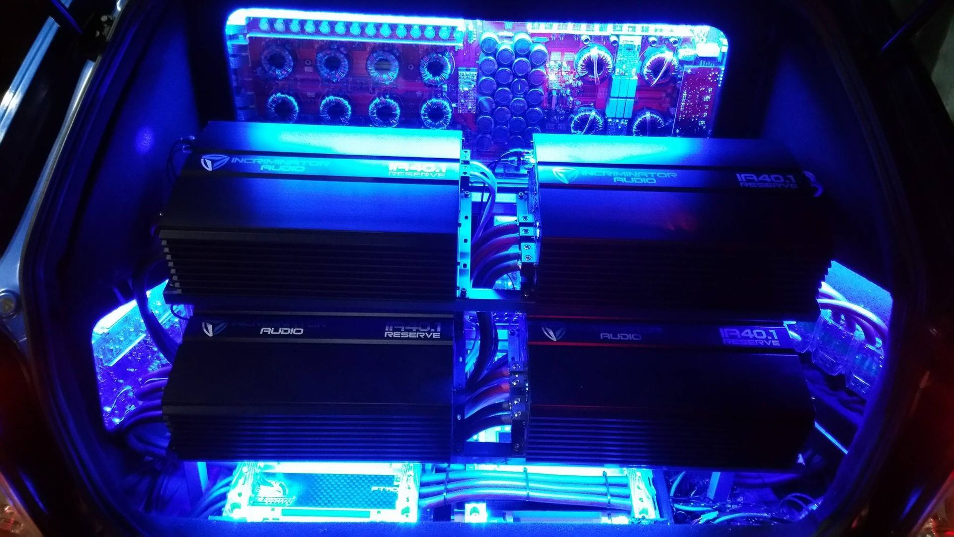 Car trunk with four amplifiers and other audio equipment, illuminated by blue lights.