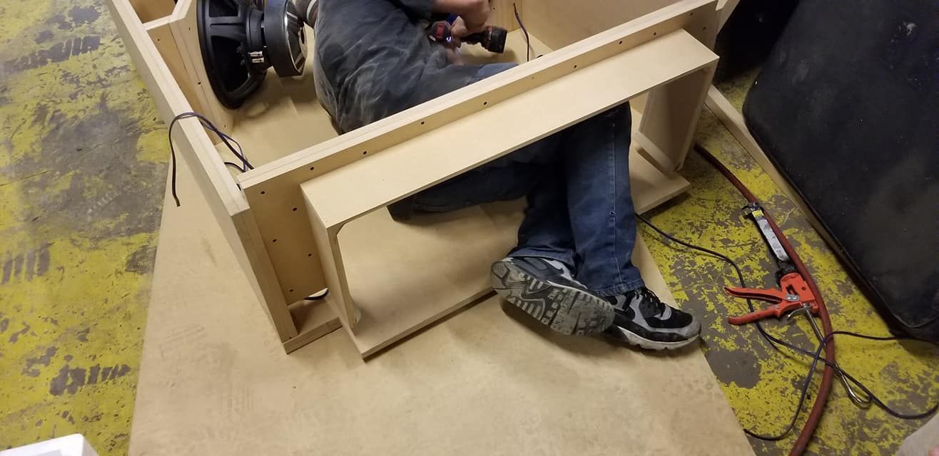 A person sits inside a wooden enclosure, likely constructing a speaker box.