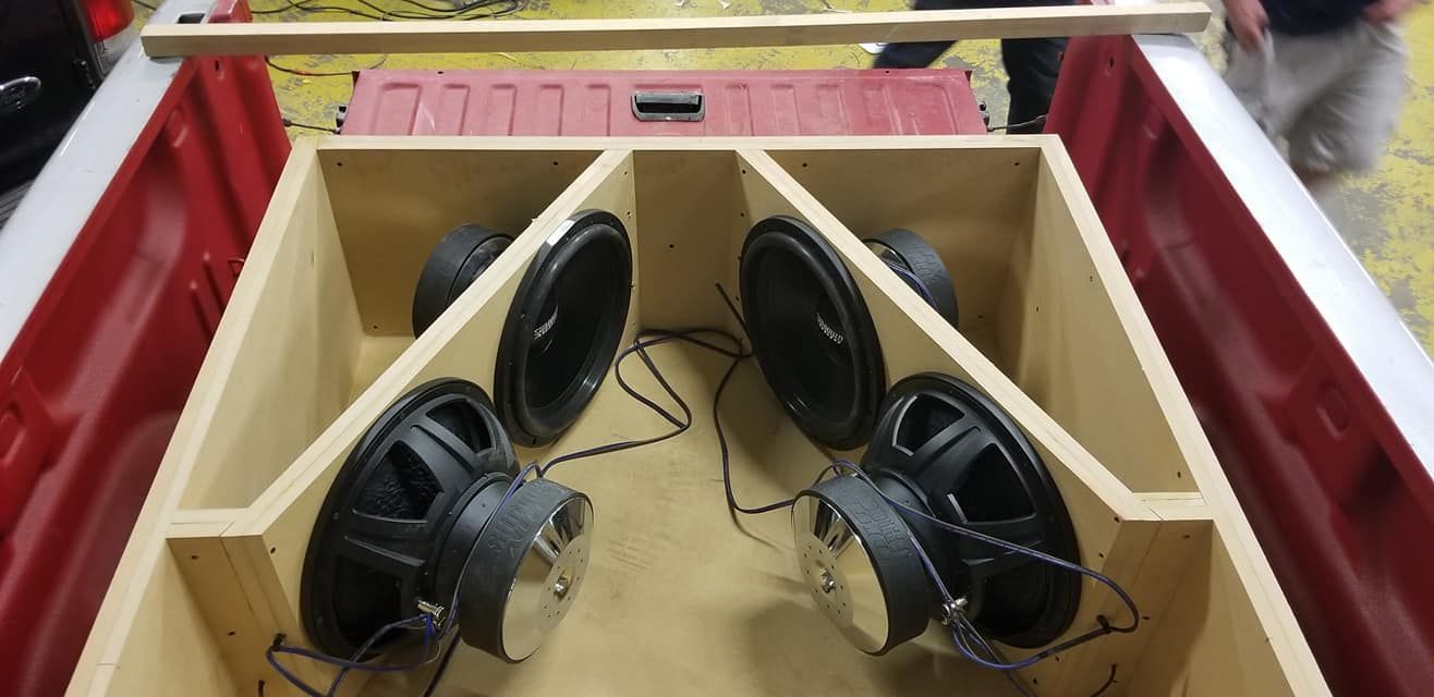 Four car subwoofers installed in a custom wooden enclosure within the bed of a red pickup truck.