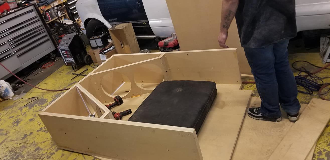 A person assembling a large, custom-built wooden subwoofer box in a garage setting. The box has multiple compartments.