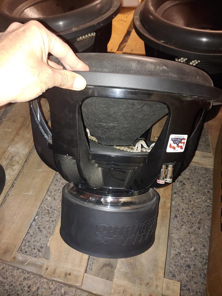Hand holding a large black subwoofer with an American flag sticker; sitting on a wooden pallet.