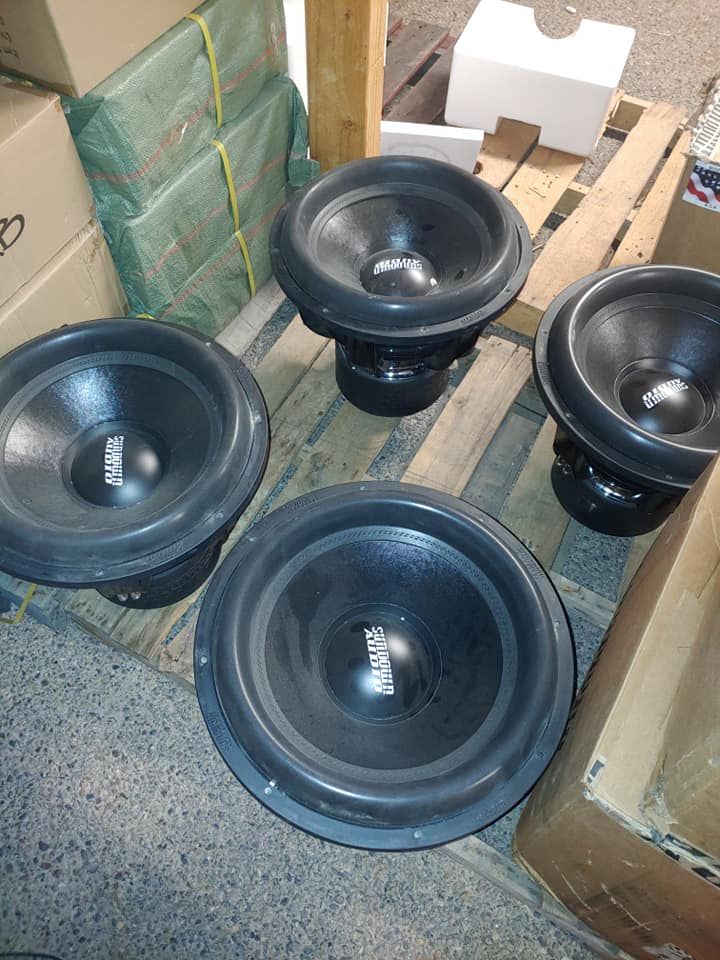 Four black subwoofer speakers arranged on a wooden pallet. The speakers are facing upward.